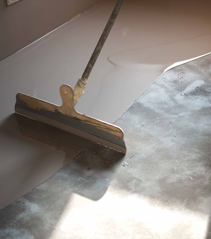 Squeegee pulling a light-colored floor coating across gray concrete in a sunlit room